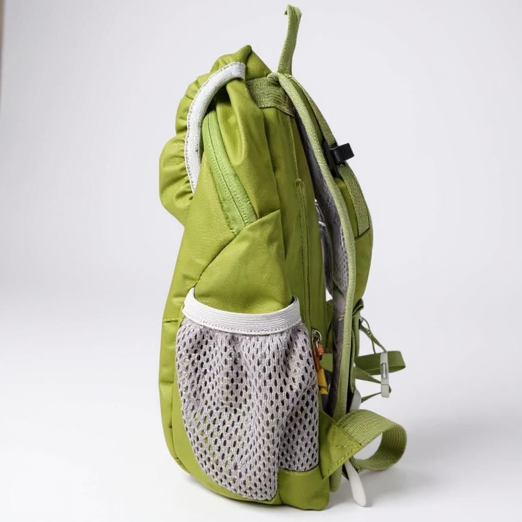 The Hiking Backpack for toddlers & kids in green, featuring gray mesh side pockets, a zipper, adjustable straps, a top handle, and space for a water bottle, stands upright on a white surface viewed from the side.
